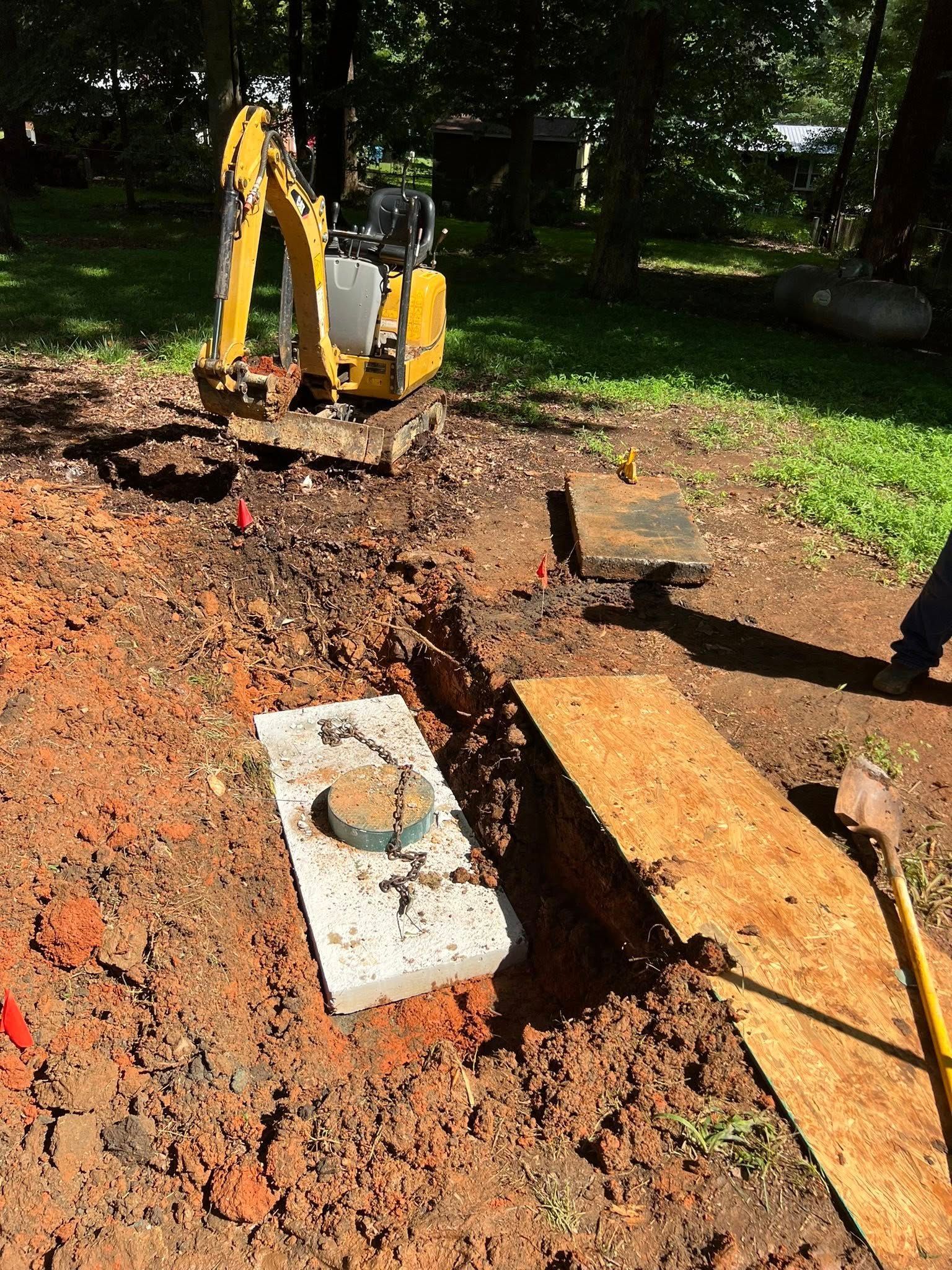 A small yellow excavator parked near a trench containing a concrete slab with a circular access cover in a wooded yard.
