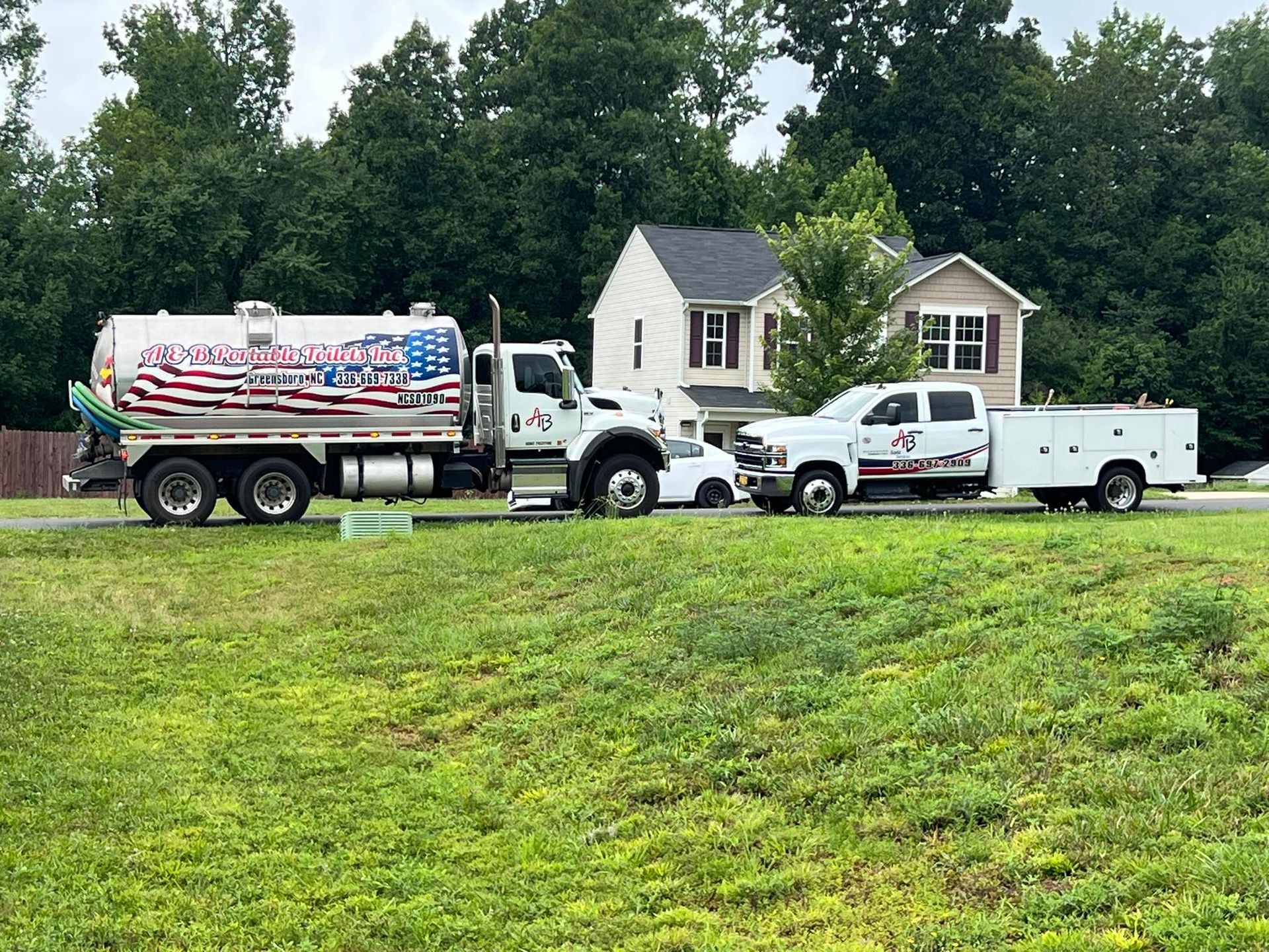 A fuel tanker truck with an American flag design and a white utility truck parked in front of a house in a grassy lot.