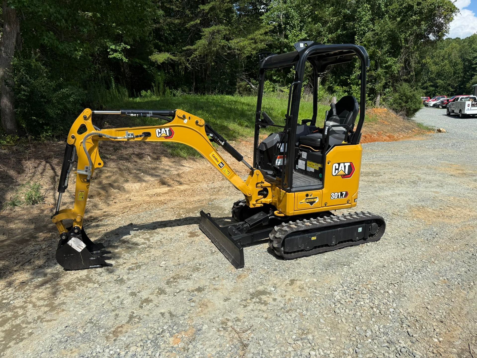 A yellow Caterpillar mini excavator parked on a gravel surface with trees in the background.