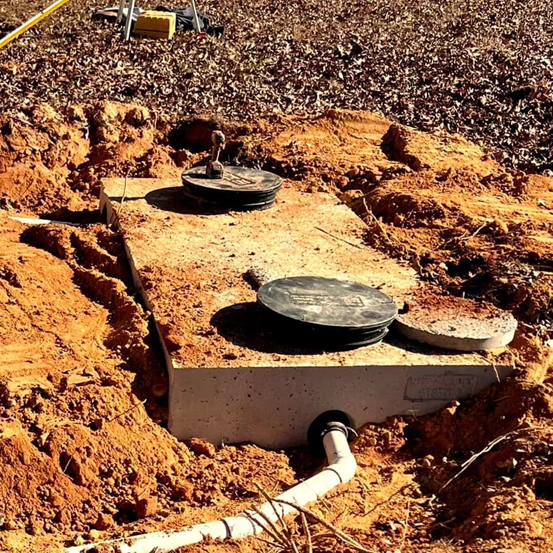 A partially buried concrete septic tank with two black lids and a connected white PVC pipe set in reddish-brown soil.