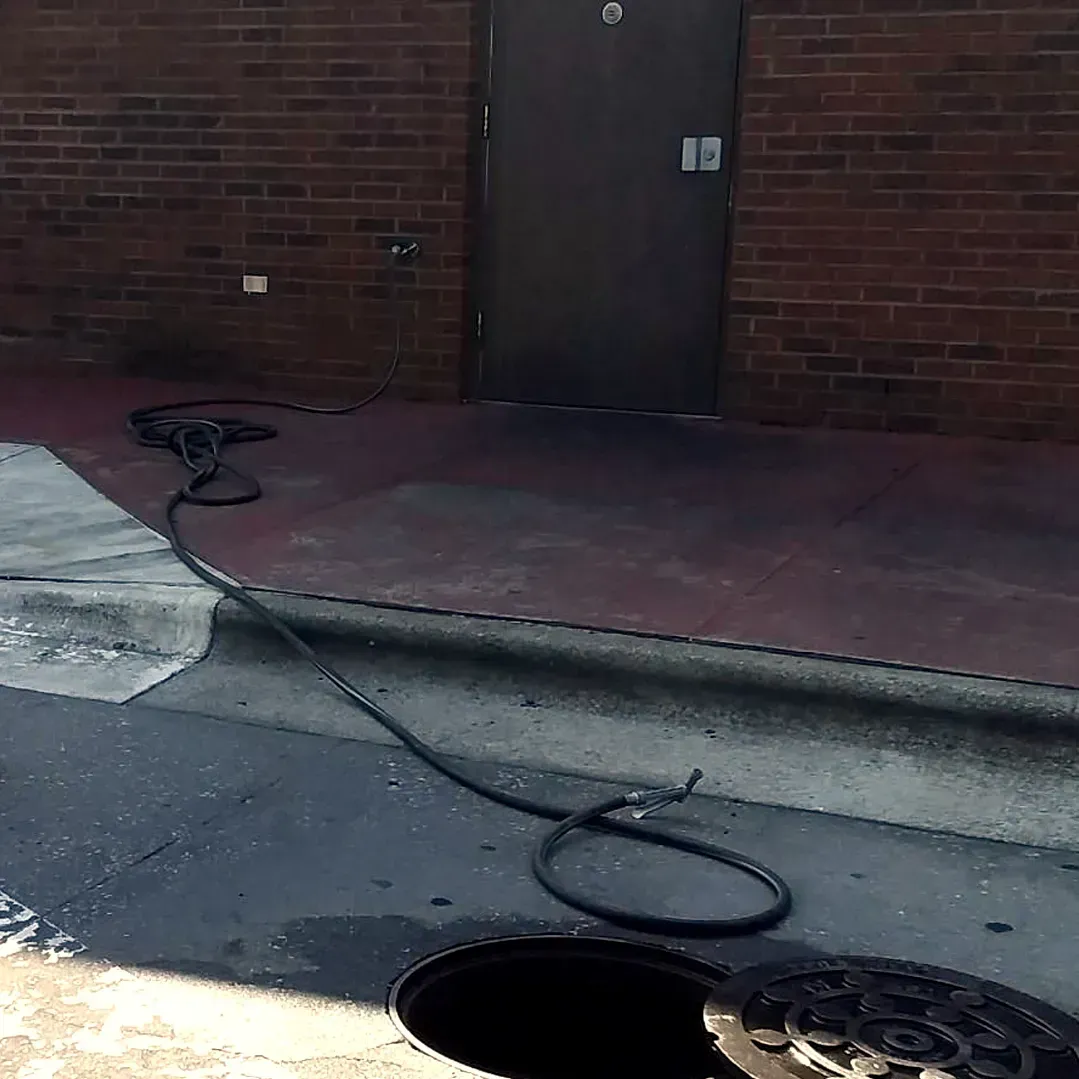 A long black hose connects a dark building wall to an open, uncovered sewer manhole in the ground.