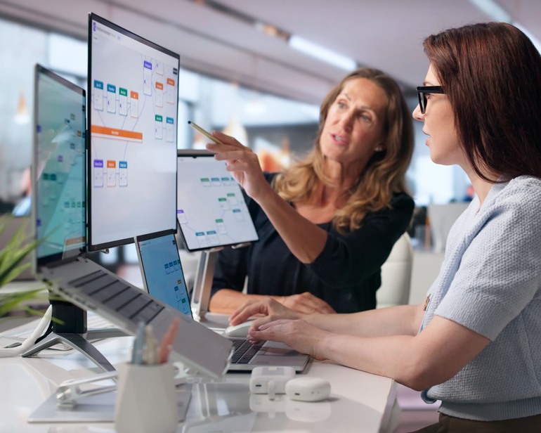 Two coworkers discussing a project at a desk with dual monitors in a bright office