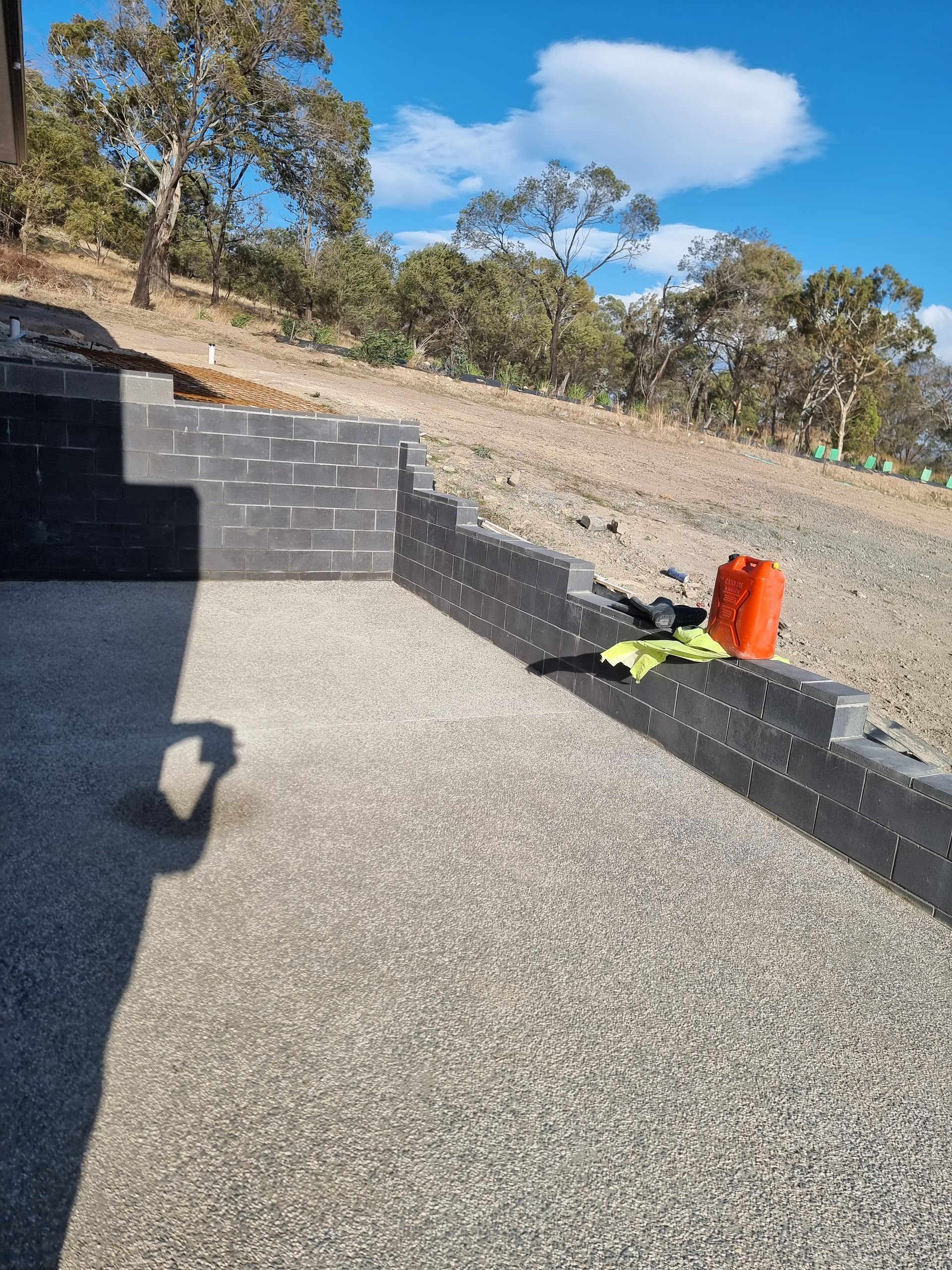 A retaining wall made of dark grey blocks under construction on a gravel patio, with trees and a blue sky in the background.