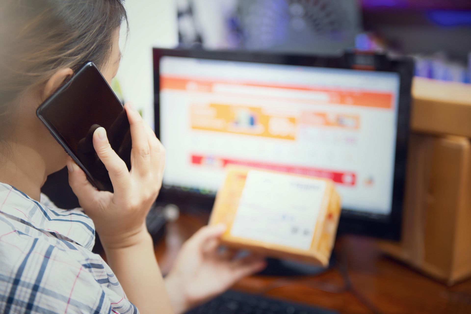 Woman on phone, holding a package, looking at a computer screen displaying an orange-themed website.