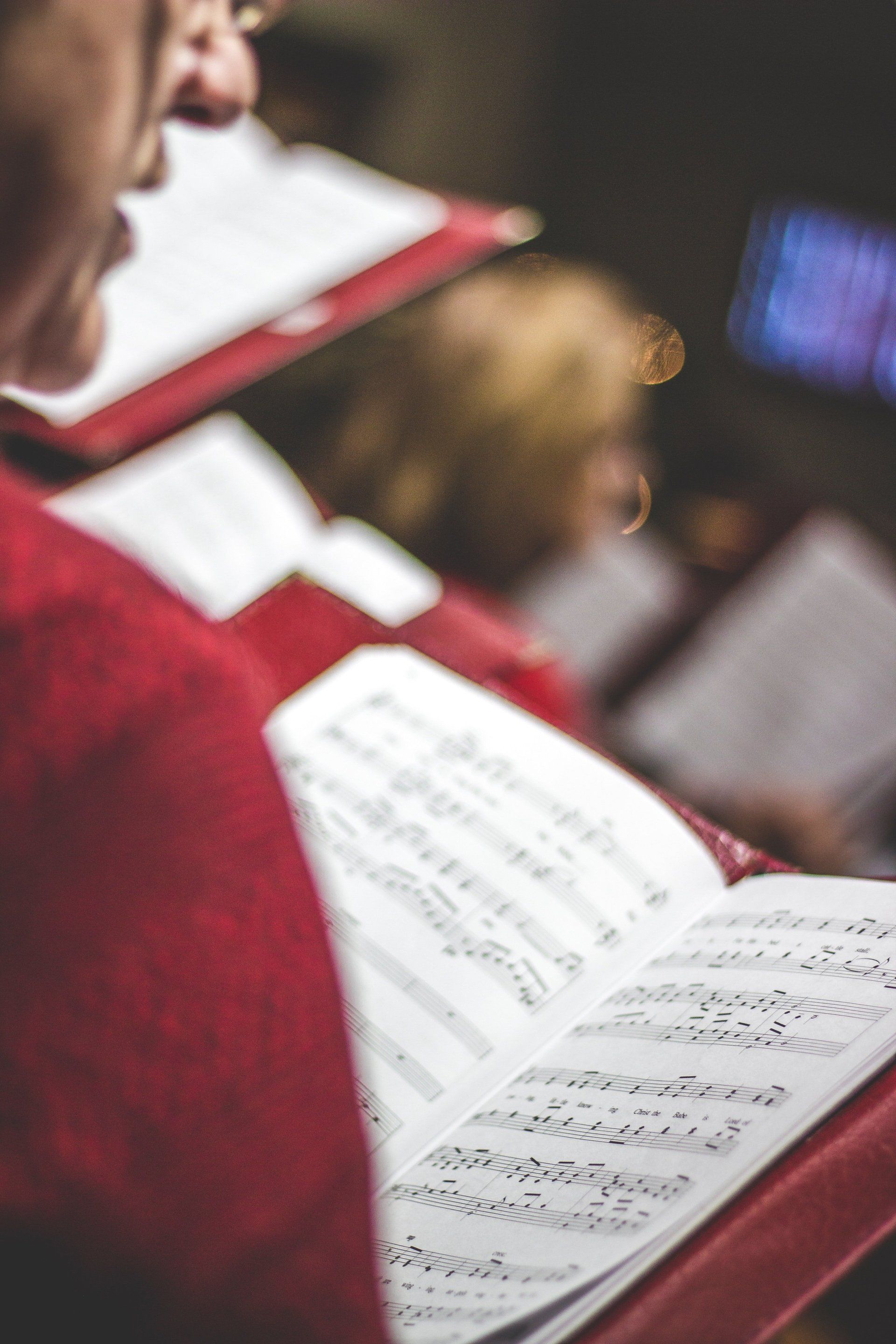 Choir members in red robes sing, reading music from open books. Blurred background, indoor setting.