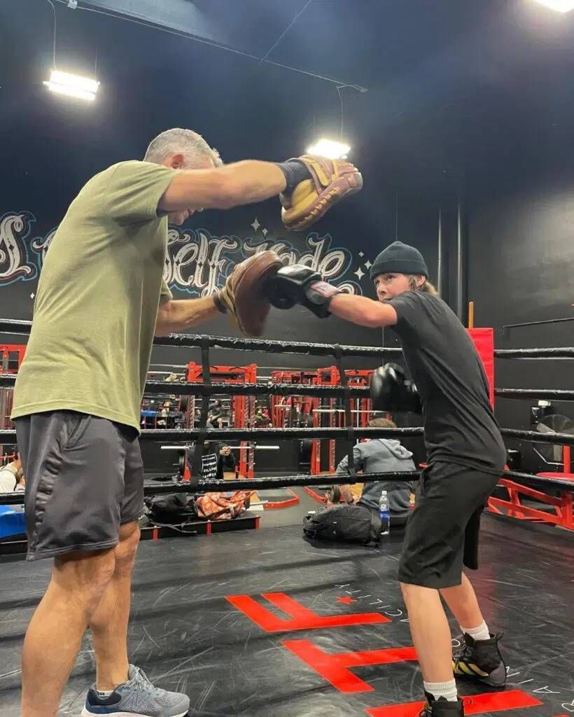 Two men sparring with boxing gloves in a gym, one holding pads, blue wall and punching bags behind them
