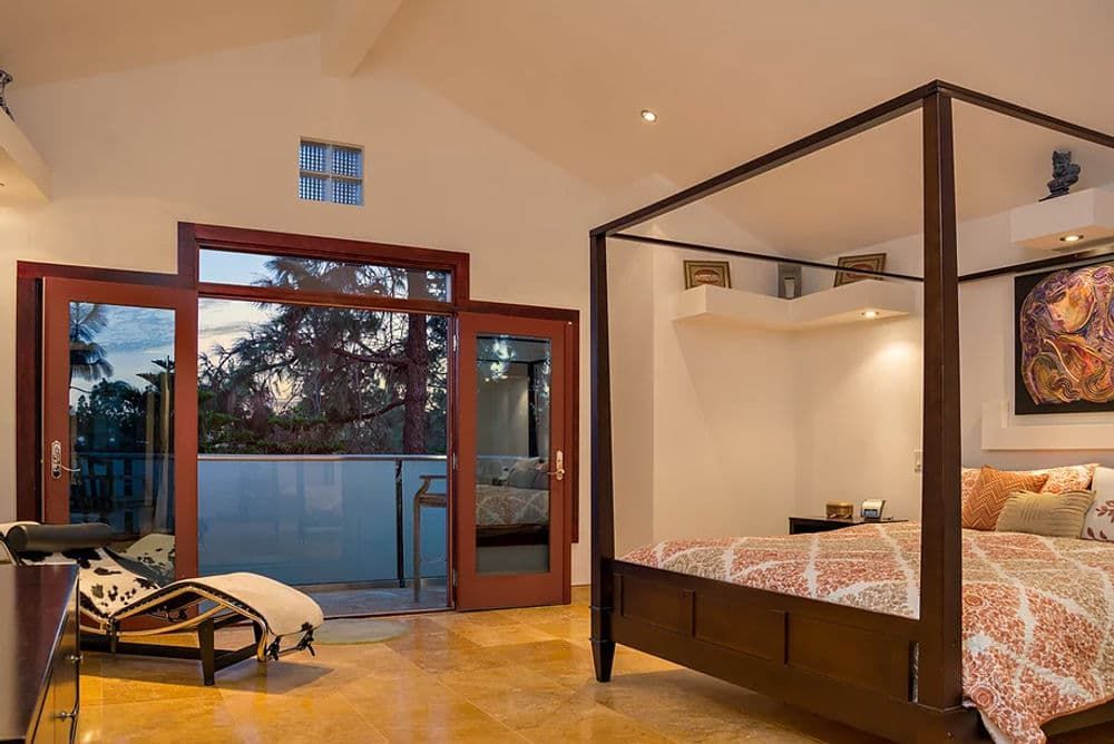 Modern bedroom with four-poster bed, open glass doors, and a balcony view at dusk