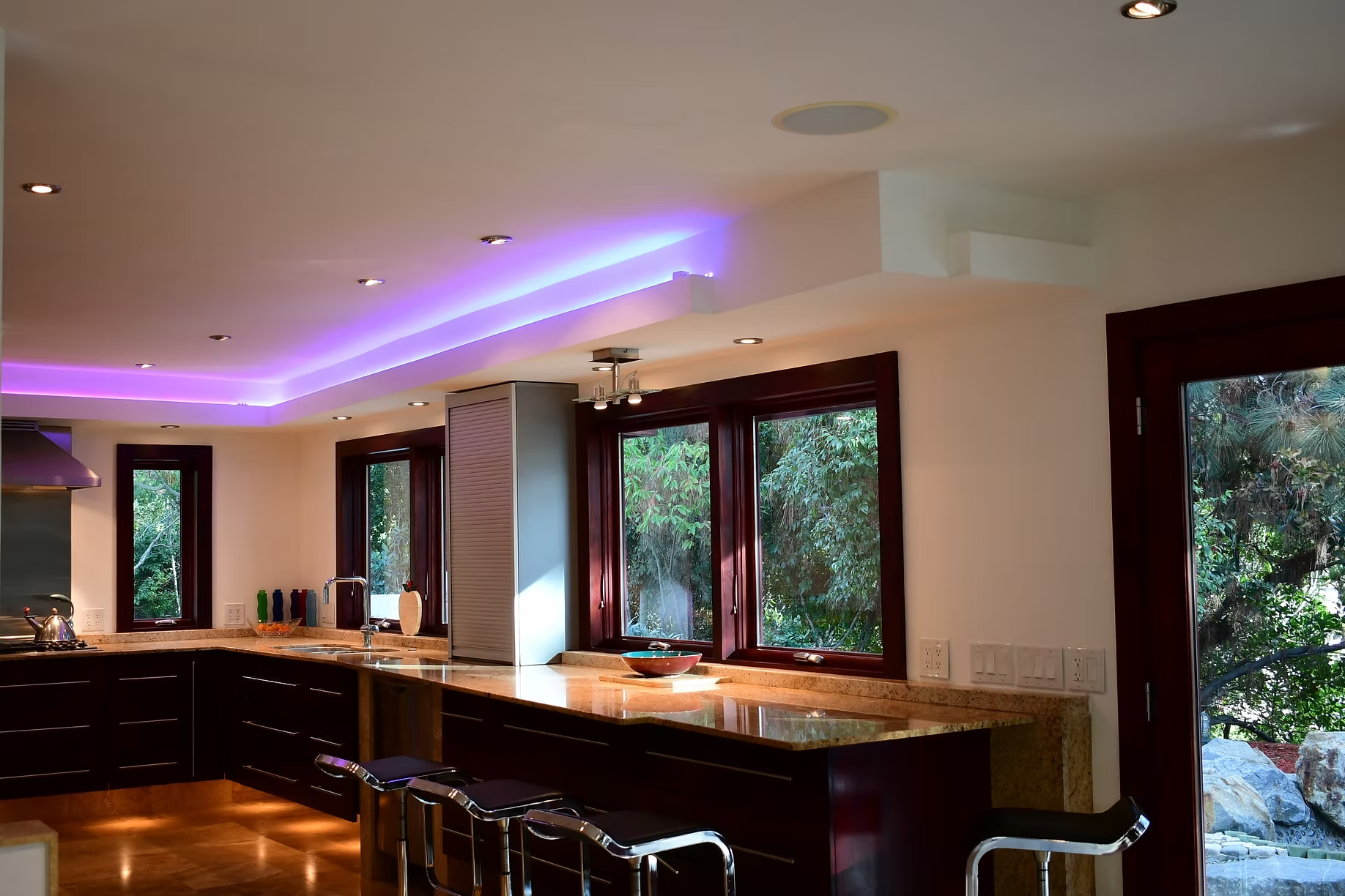 Modern kitchen with dark wood cabinets, marble island, and purple accent lighting by large windows