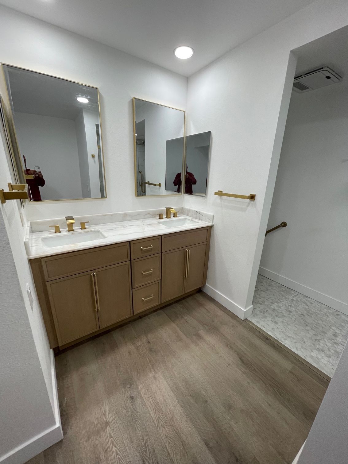 Modern bathroom with double vanity, large mirrors, tan cabinets, and gray wood-look flooring.