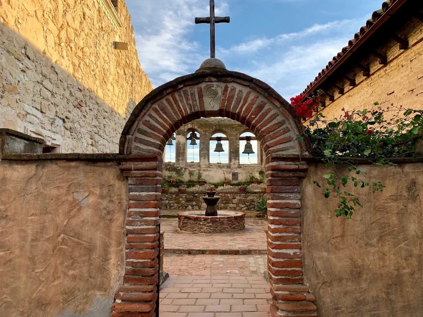 Stone archway with a cross and bells framing a small courtyard fountain between rustic walls.