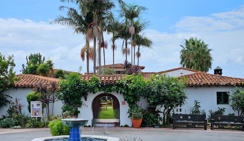 Spanish-style white house with red tile roof, arched entrance, palm trees, and lush greenery under a blue sky