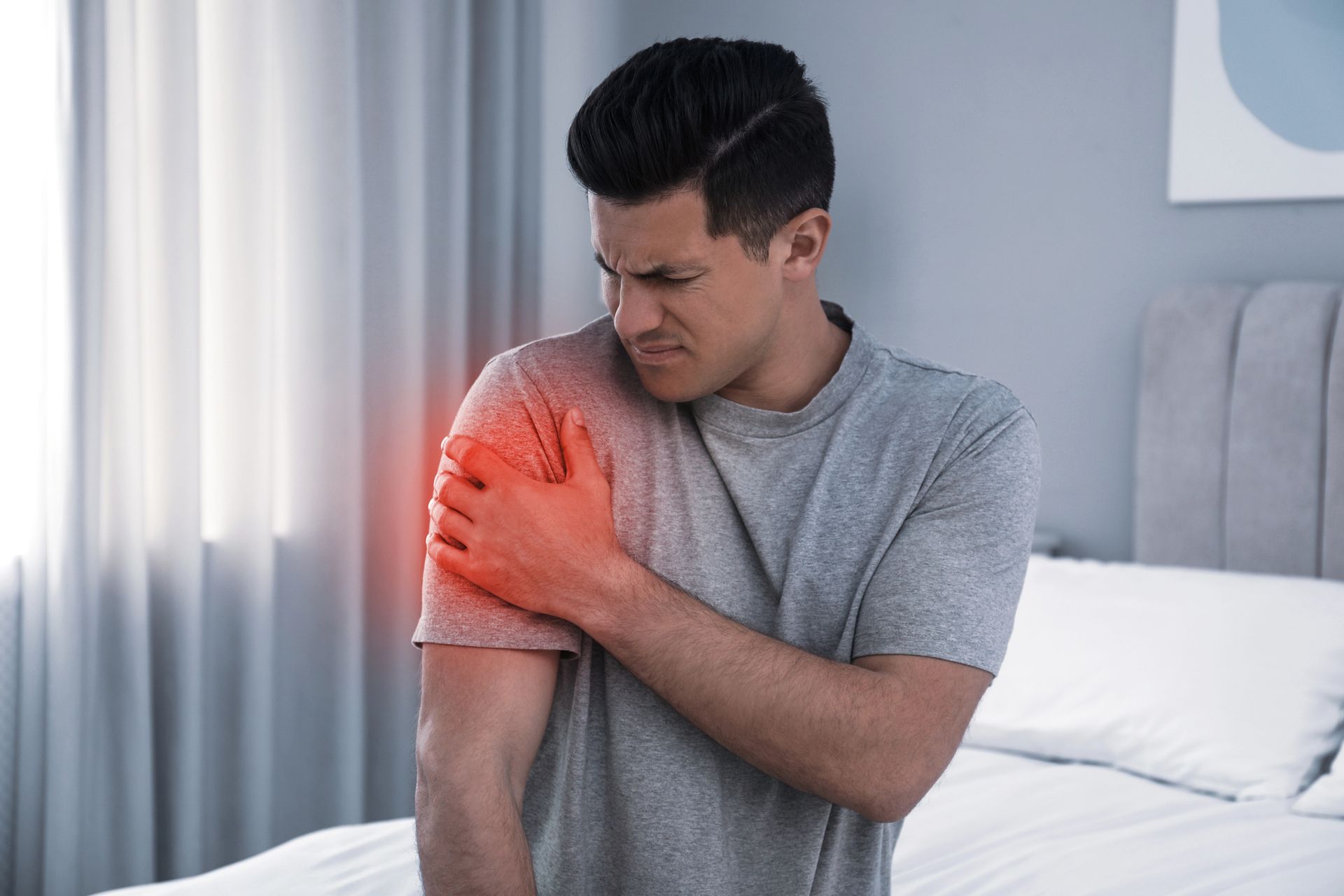 Man clutching his shoulder, indicated with red highlight, showing pain. Bedroom setting.