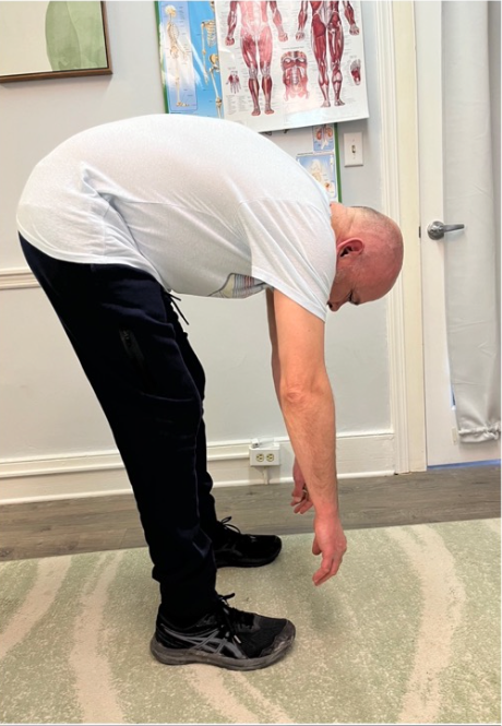 Person bending forward with arms hanging down in a clinic room, near an anatomy poster.