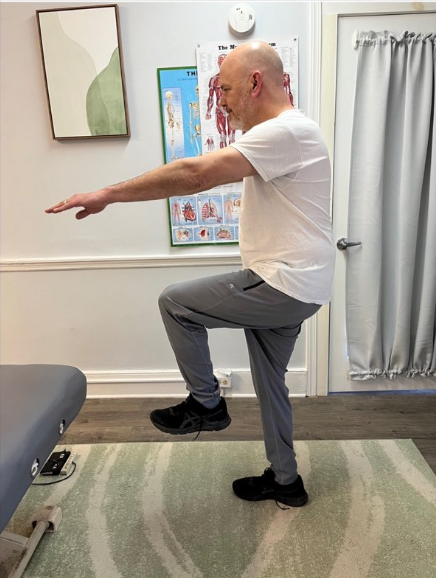 Person balancing on one leg with arms extended in a clinic therapy room.