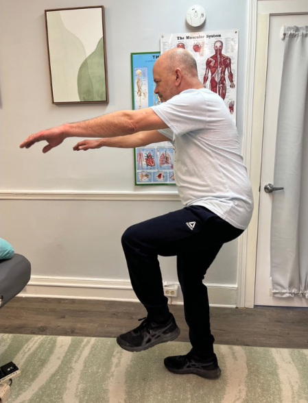 Person balancing on one leg with arms extended in a clinic room, demonstrating a leg exercise.