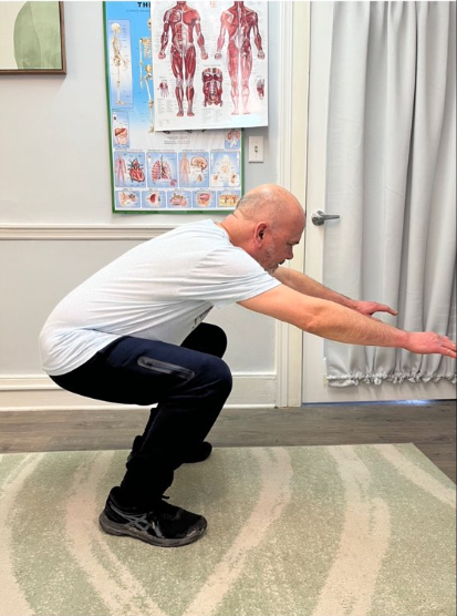 Man squats with arms extended forward in a therapy room, demonstrating a balance exercise.