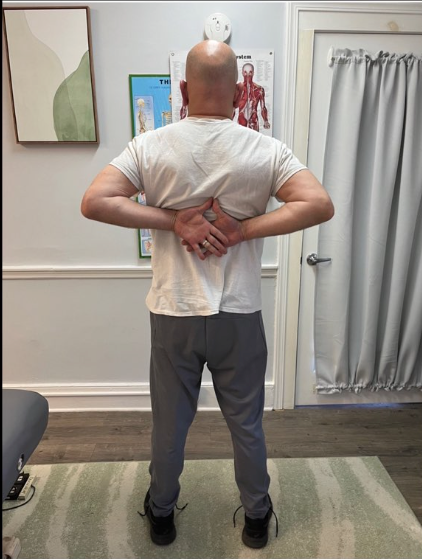 Person standing in a clinic, facing away, hands pressed behind the lower back during a posture assessment.