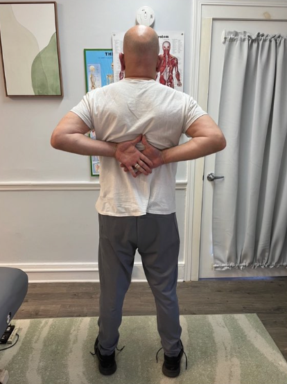 Person standing in a clinic, reaching both hands behind their back during a posture assessment.