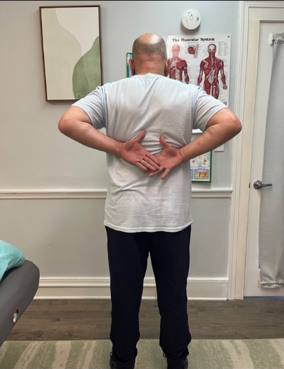 Person standing with hands on lower back in a clinic exam room, facing anatomical posters.