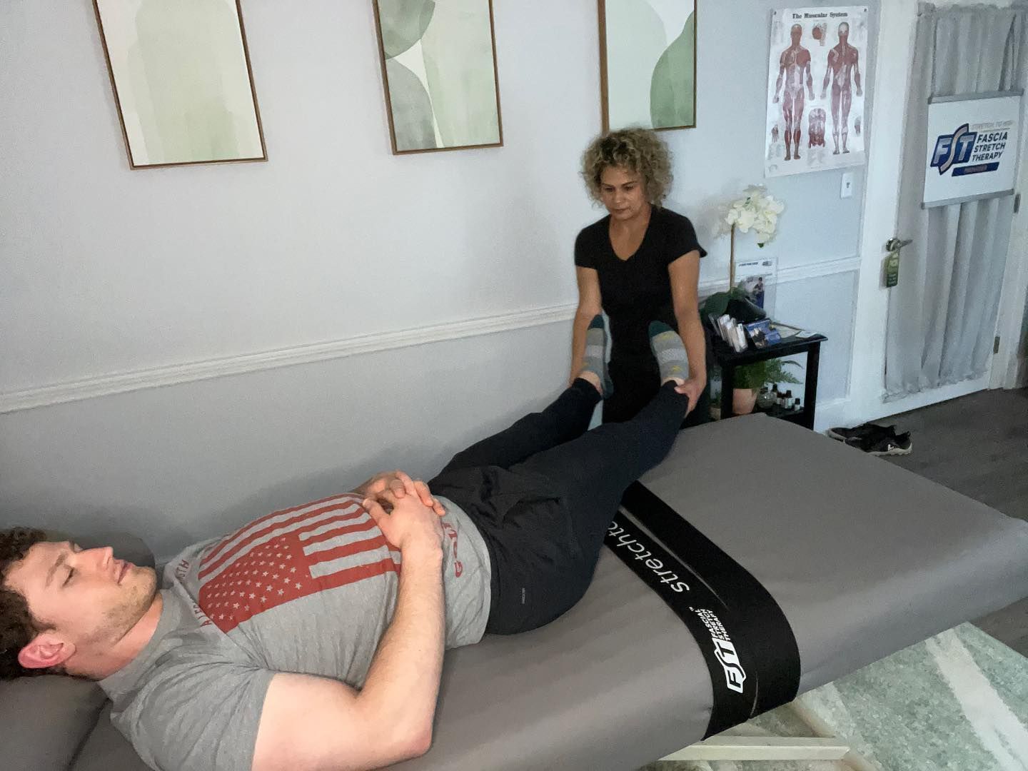 Person lies on a massage table while a therapist stretches their leg in a treatment room.