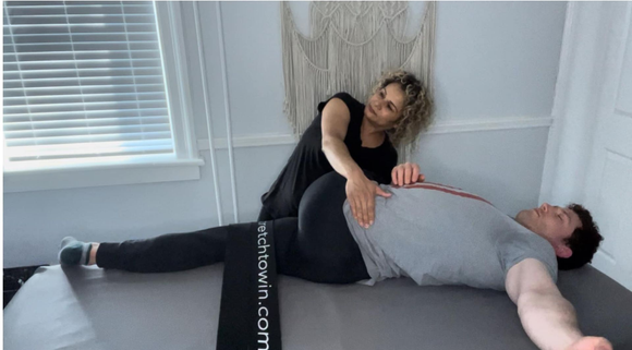 Two people on a mat, one performing a side stretch while holding the other’s torso, in a bright room
