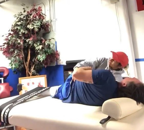 Person in red cap gently stretching another person on a white massage table in a bright therapy room