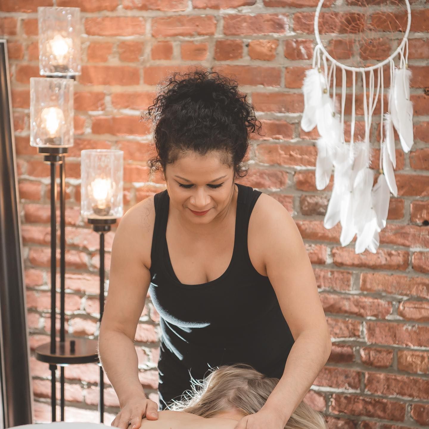 Woman in black tank top styling hair in front of a brick wall and dreamcatcher decor