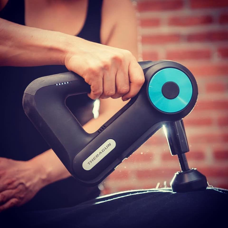 Person using a black and teal massage gun on a shoulder, with a brick wall background