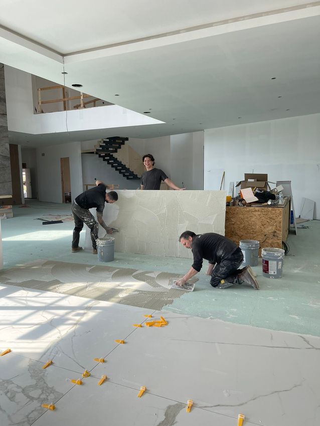 Three workers laying tile in a room under construction. One works on a decorative counter. Bright, open space.