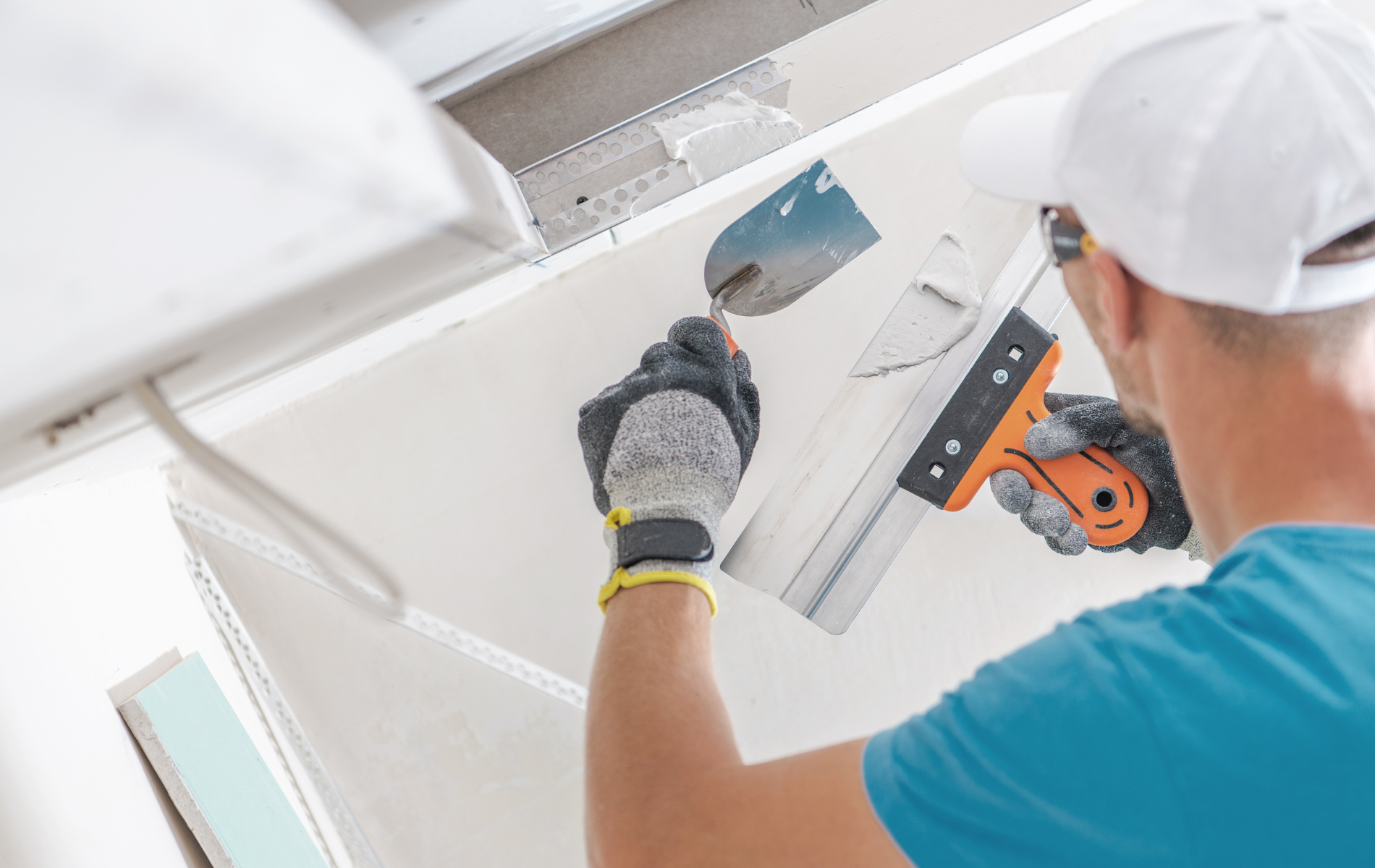 Person in a blue shirt and white hat applying joint compound to drywall with two trowels.