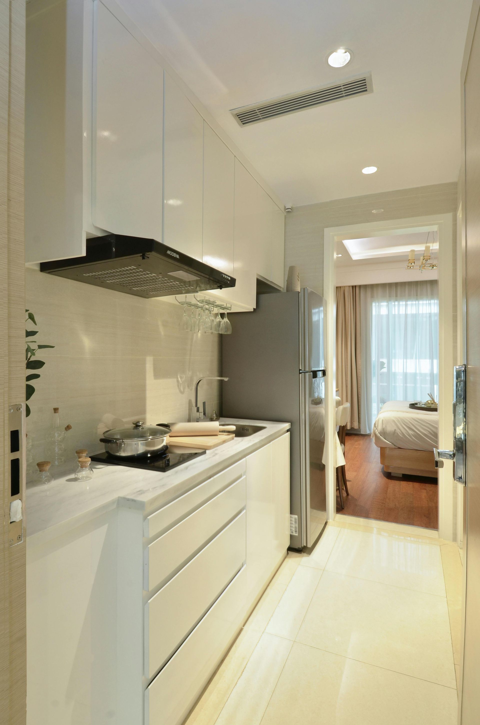 Narrow white kitchen with stainless steel appliances, leading to a bedroom.
