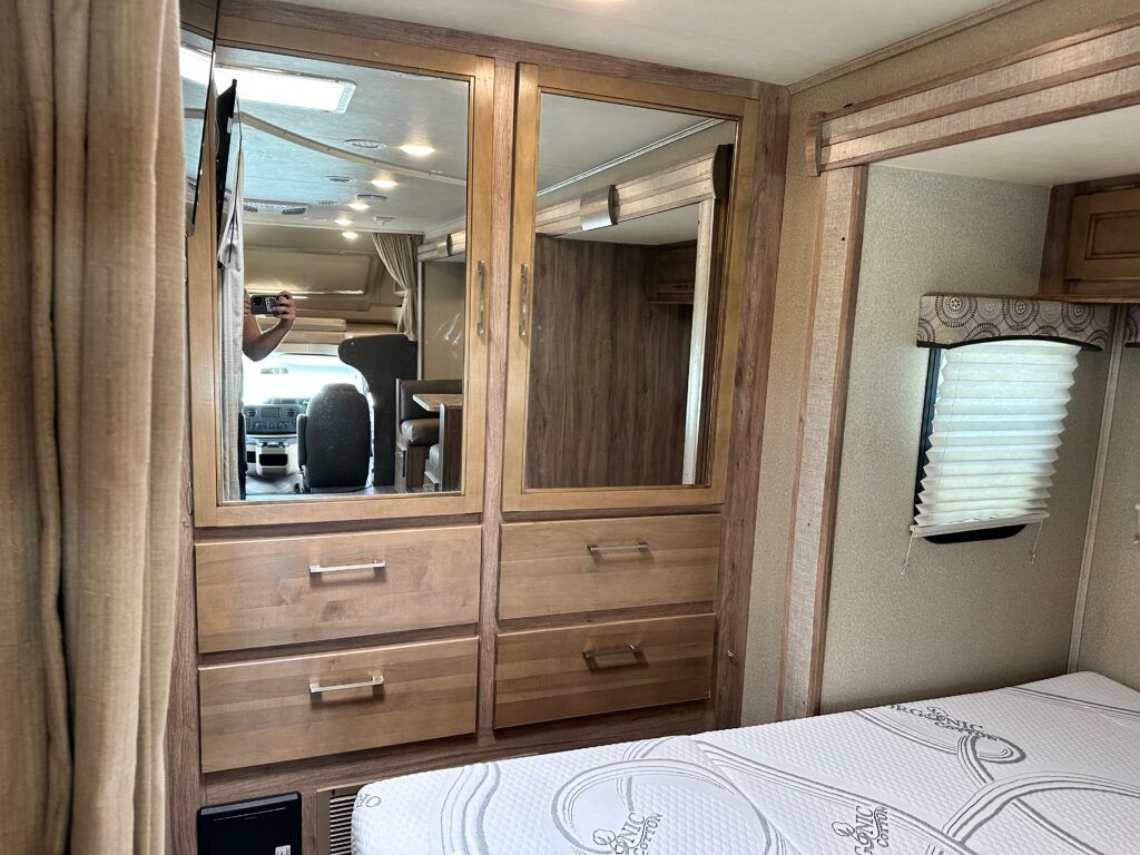 A bedroom in a rv with a bed , dresser , mirror and drawers.