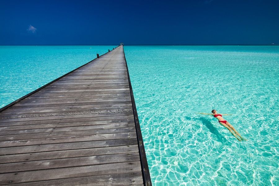 Wooden pier stretching into turquoise water, with a swimmer floating beside it under a clear blue sky.