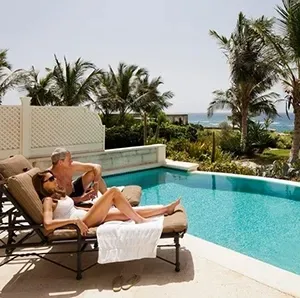 Couple relaxing on lounge chairs beside a turquoise pool with palm trees and ocean view