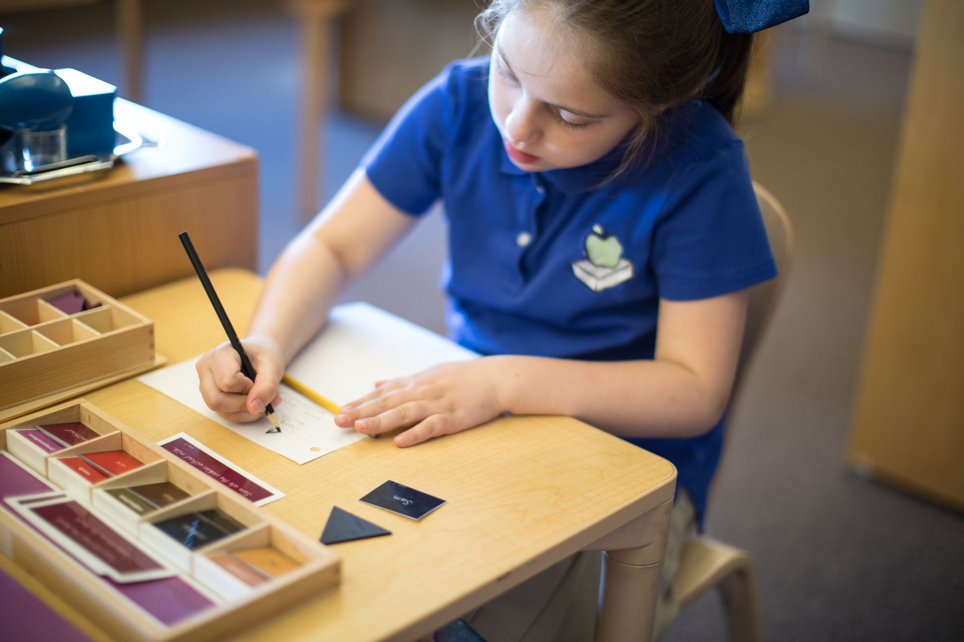 Student in blue shirt writing on paper at a classroom table with learning materials nearby