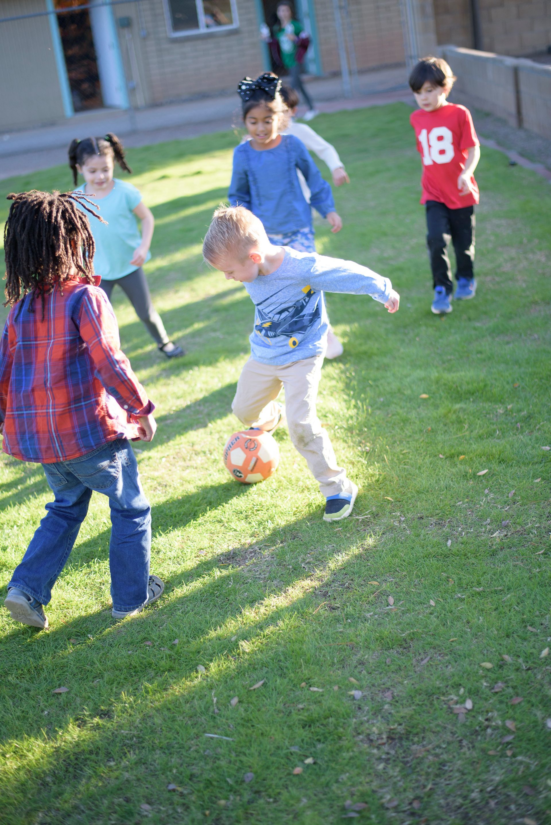 Children playing soccer on a grassy field near a building, with one kicking an orange ball.