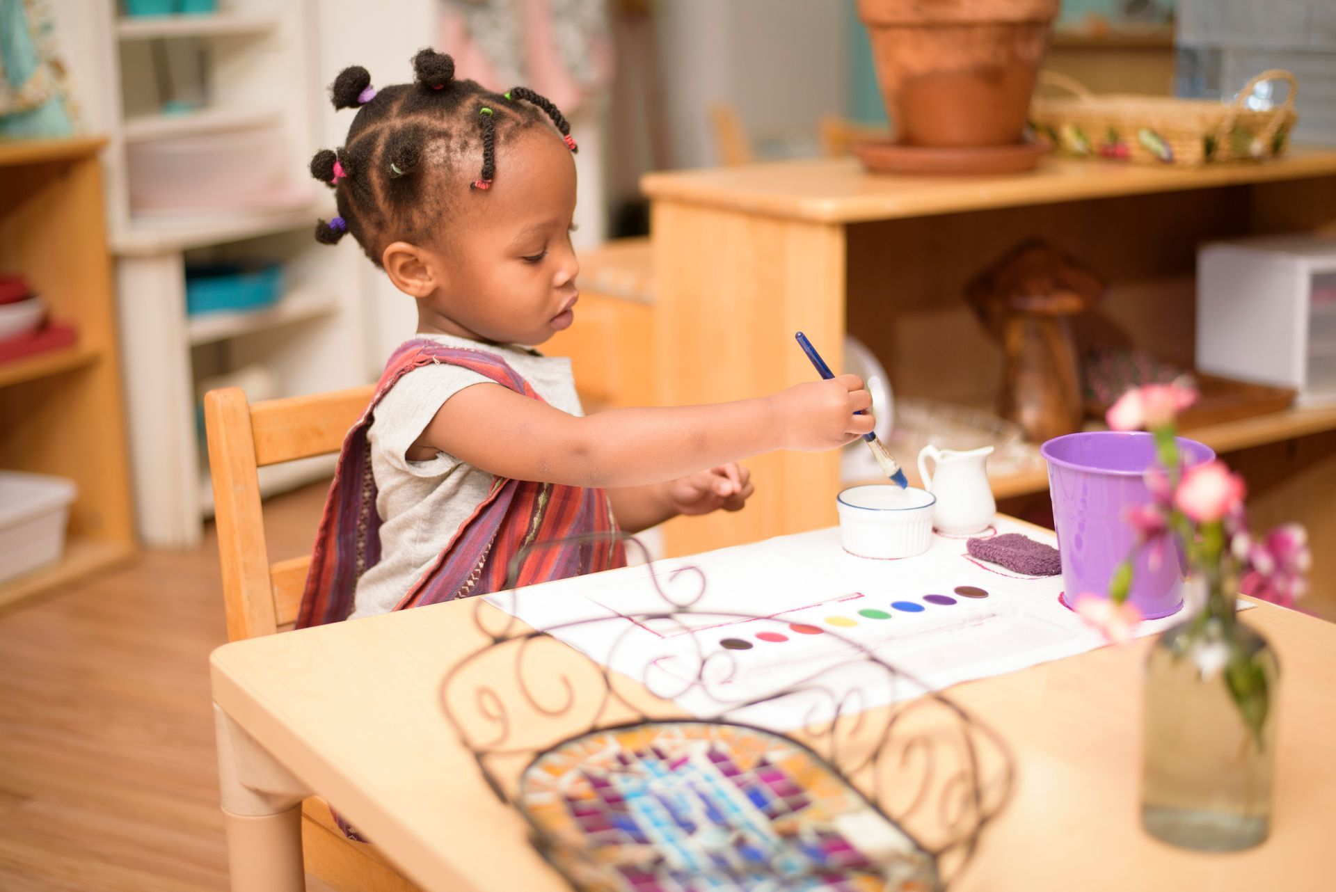 a Montessori little girl is sitting at a table painting with a brush .