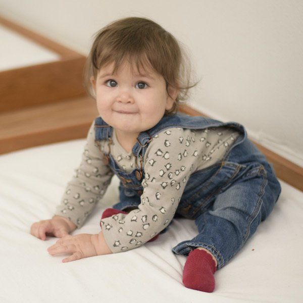 a Montessori baby wearing overalls and red socks is sitting on a bed