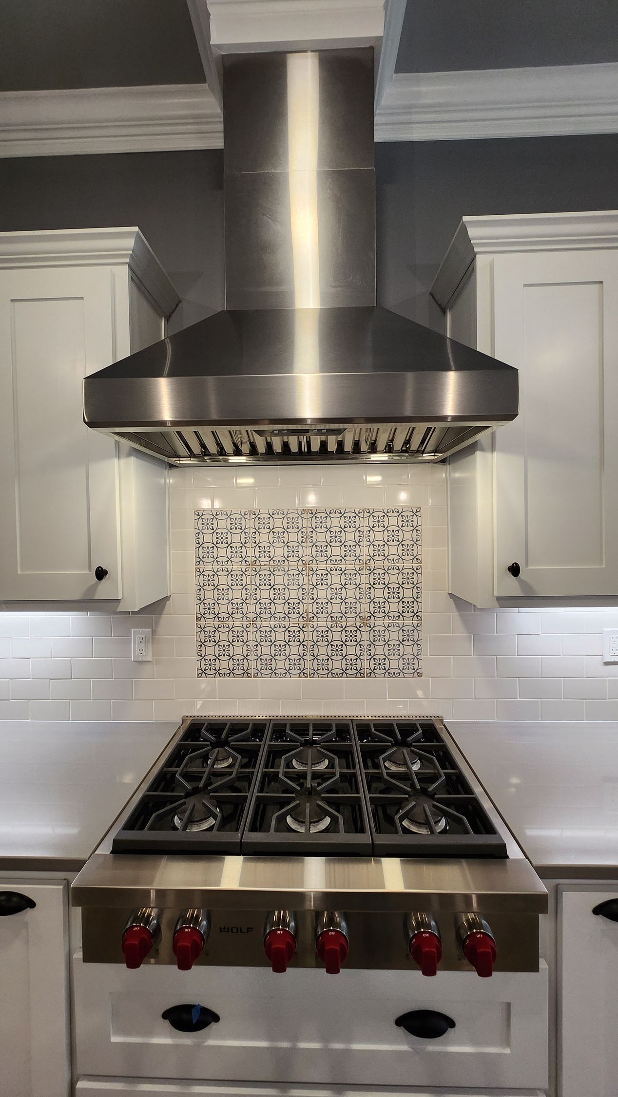 Stainless steel range hood above a gas range in a white kitchen with decorative tile backsplash.