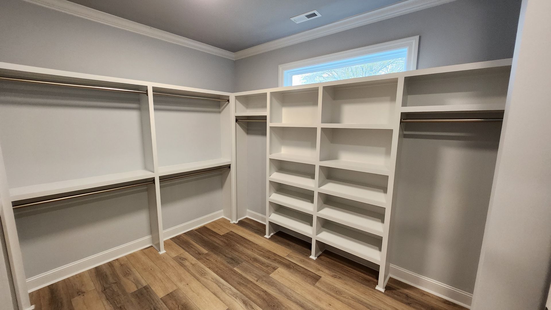 Empty walk-in closet with white shelving and hanging rods. Gray walls, wood-look floor, window at the back.