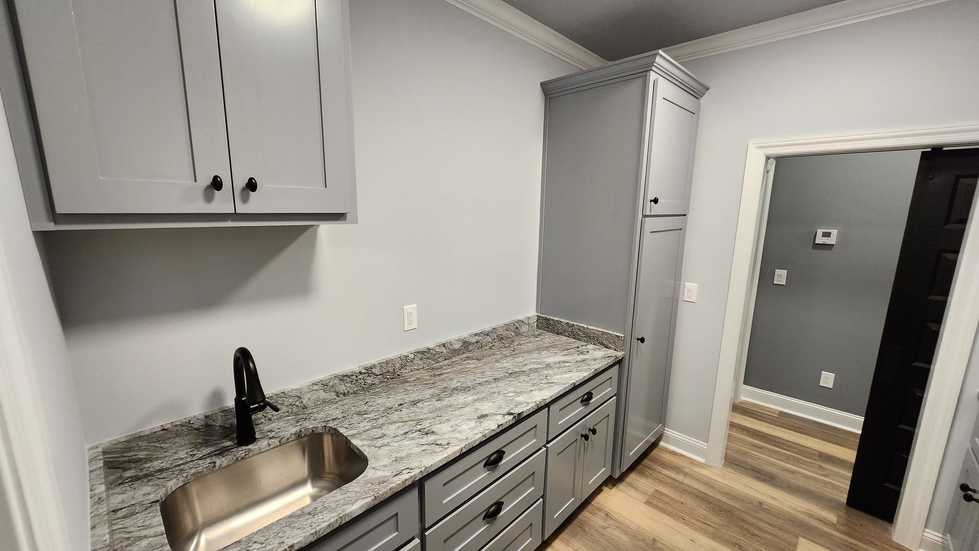 A utility room with grey cabinets, granite countertop, sink, and tall storage cabinet. Doorway leads to another room.