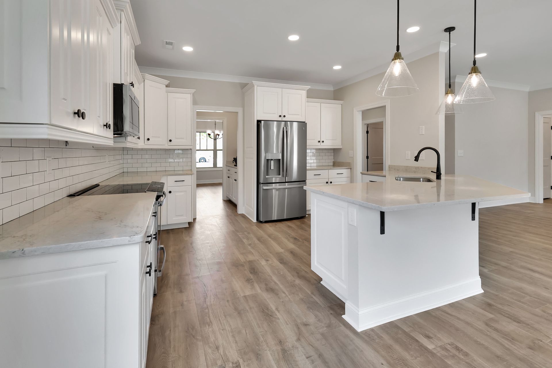 White kitchen with island, stainless steel fridge, and pendant lights. Light wood-look flooring.