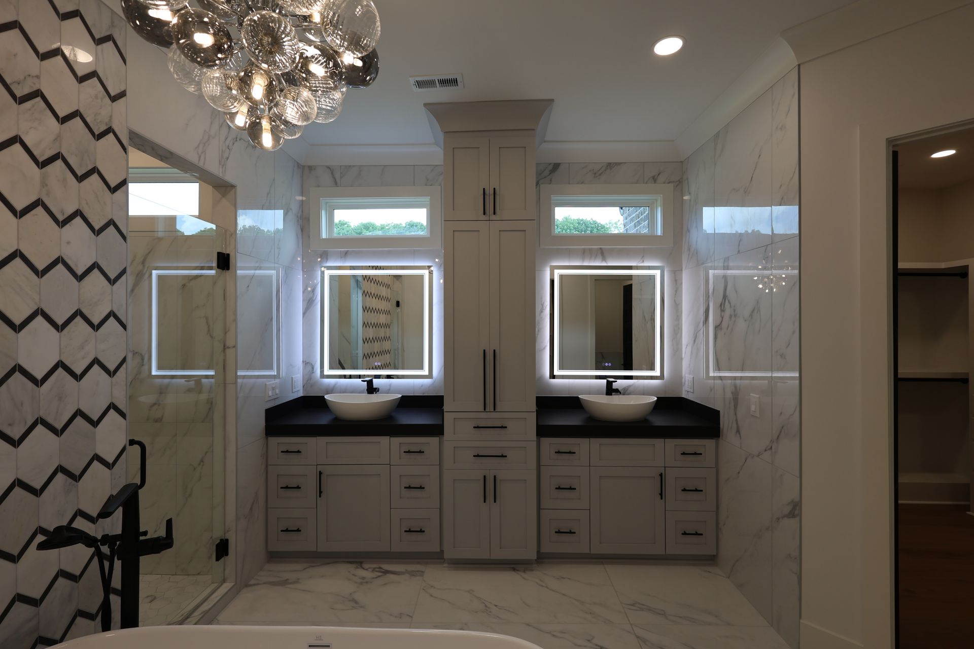 Modern bathroom with marble walls and floor, two sinks with lit mirrors, a central cabinet, and a zigzag accent wall.