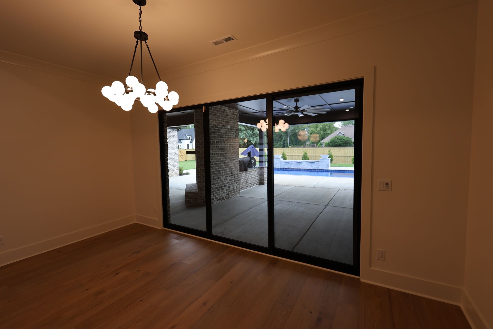 Empty dining room with sliding glass doors overlooking a patio and pool. Chandelier hangs above wooden floor.