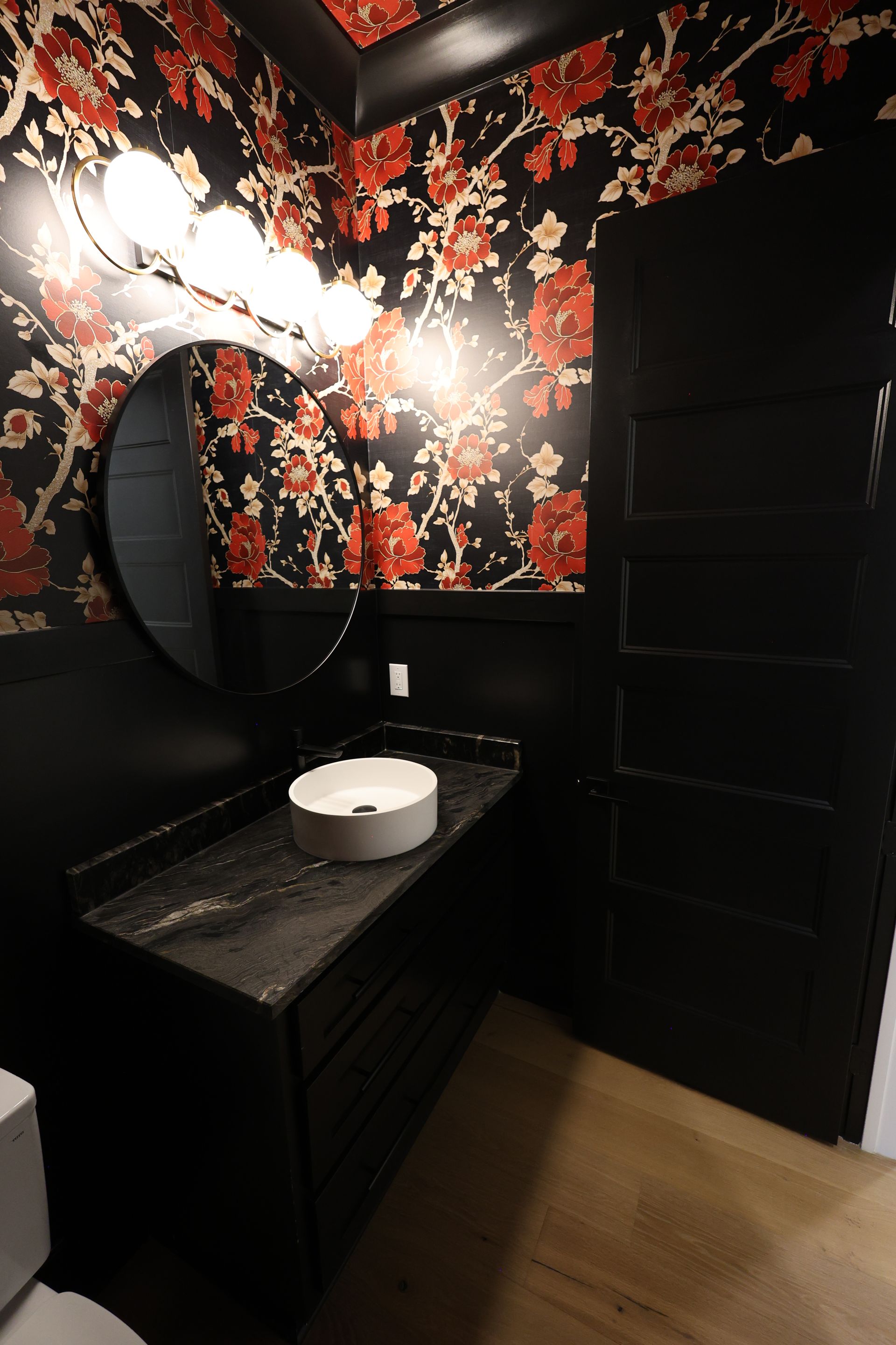 Black powder room with floral wallpaper, black vanity, and round mirror.