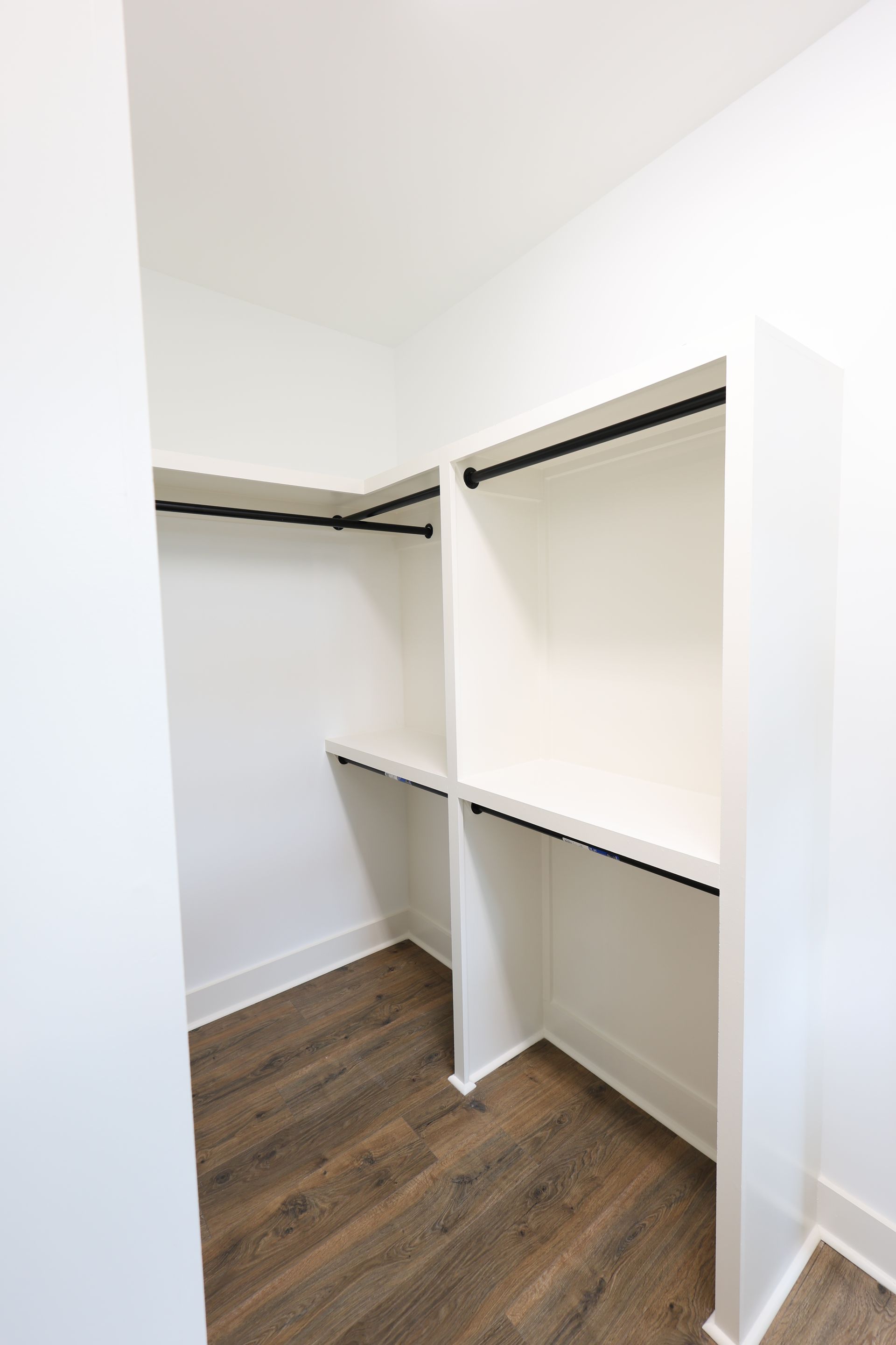 Empty white closet with shelving and hanging rods; dark wood-look flooring.