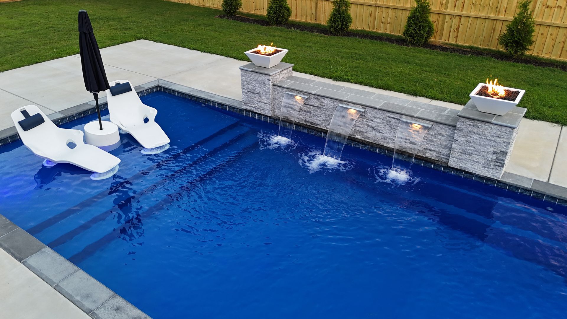Swimming pool with waterfall feature, lounge chairs, and fire pit. Blue water and gray stone accents.