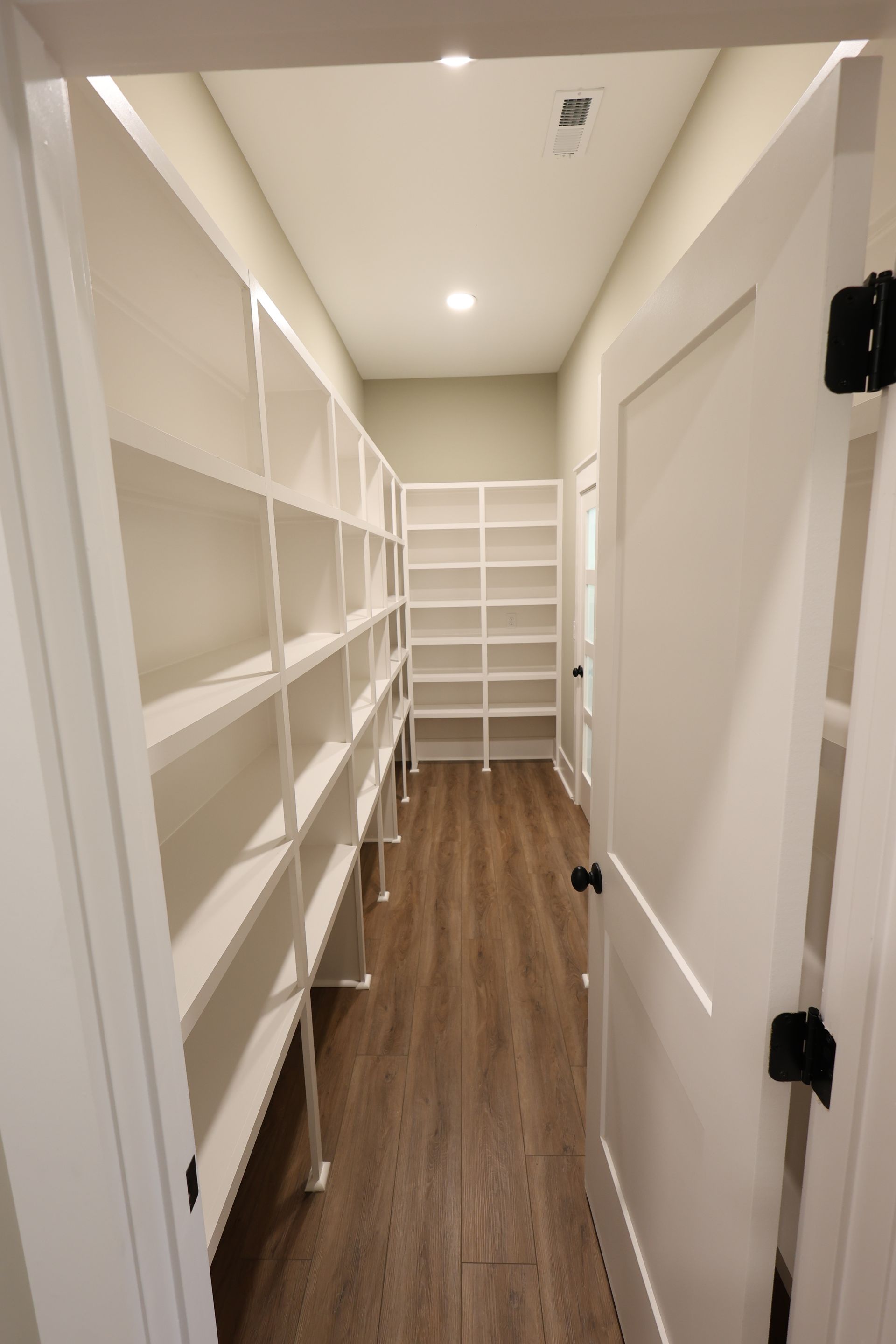 White shelves line a pantry hallway with a wooden floor. A white door is on the right.