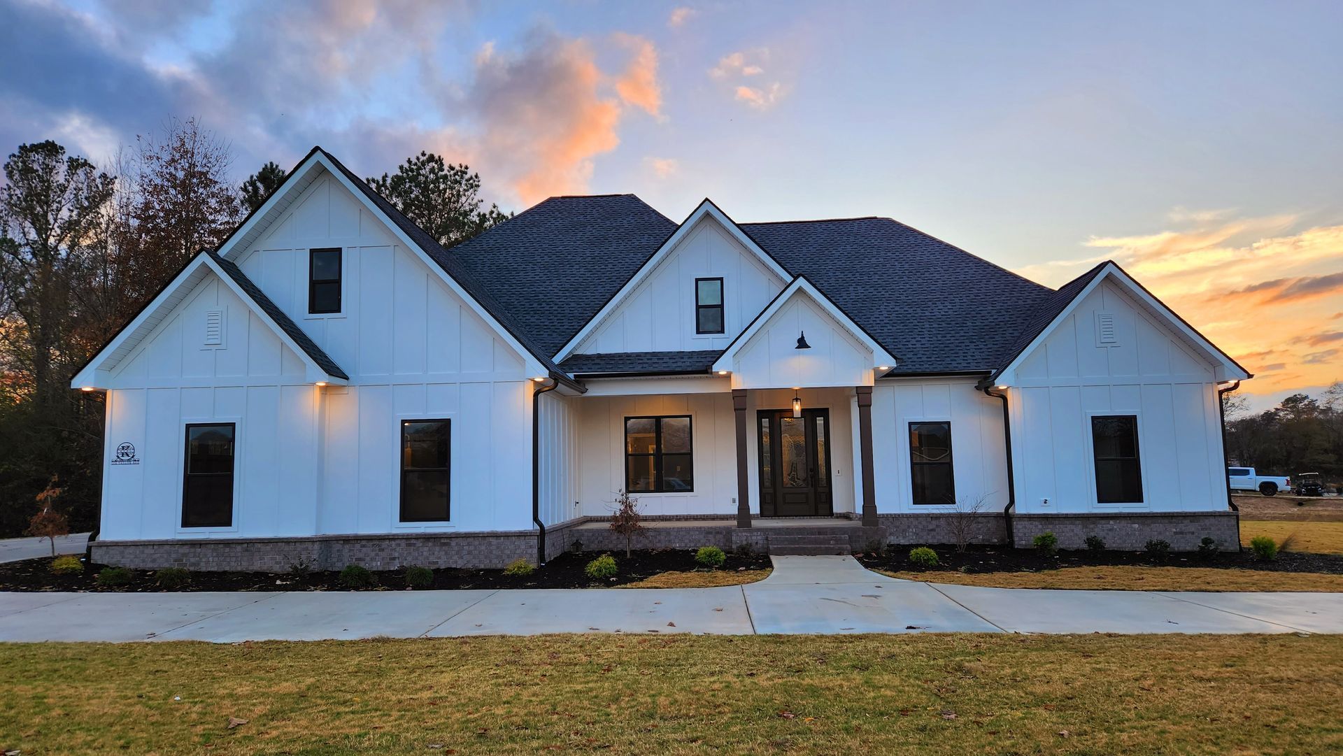 White house with dark roof and trim, brick base, set against a sunset sky.