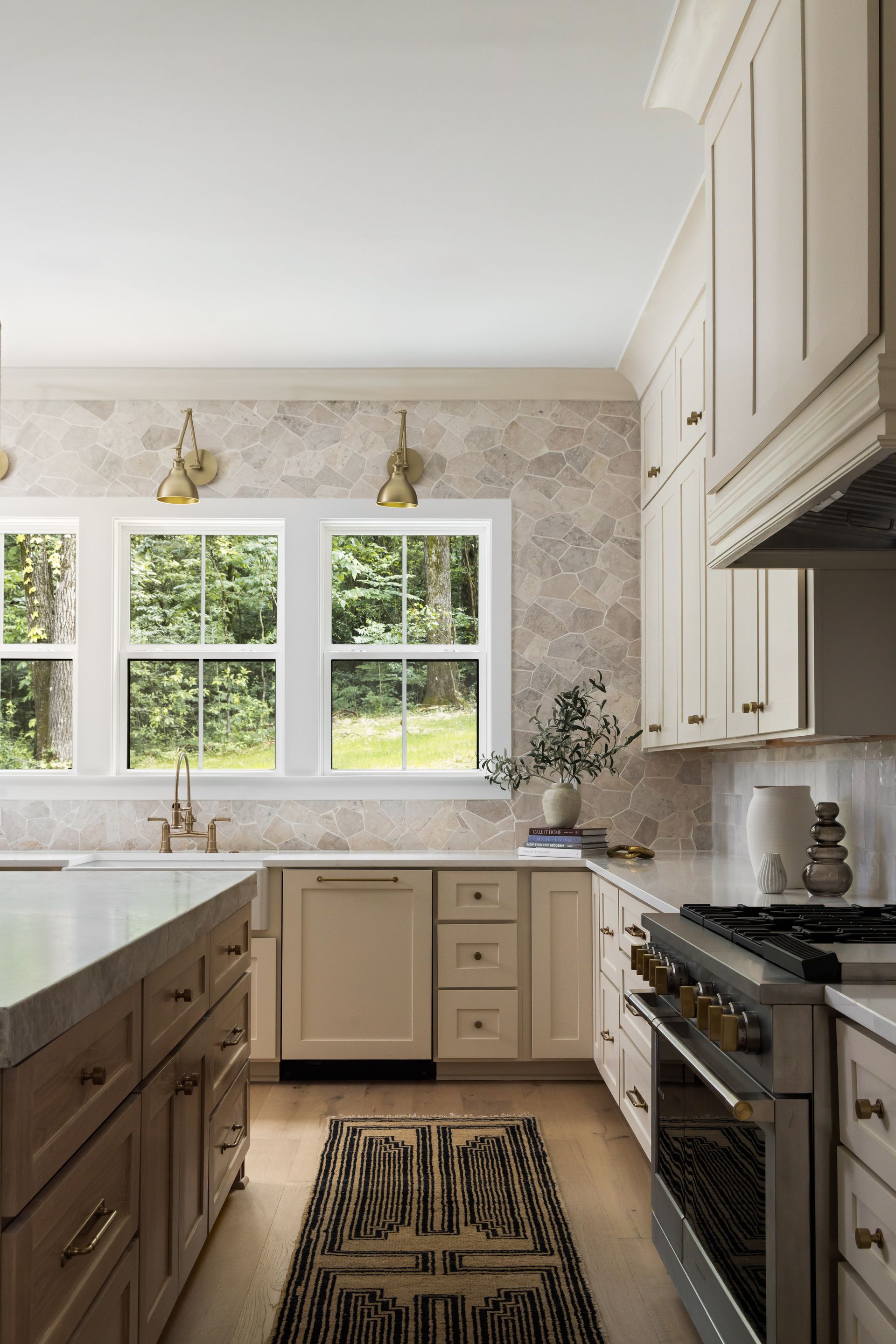 Cream-colored kitchen with island and cabinetry. Windows overlook greenery, neutral patterned backsplash, and rug.